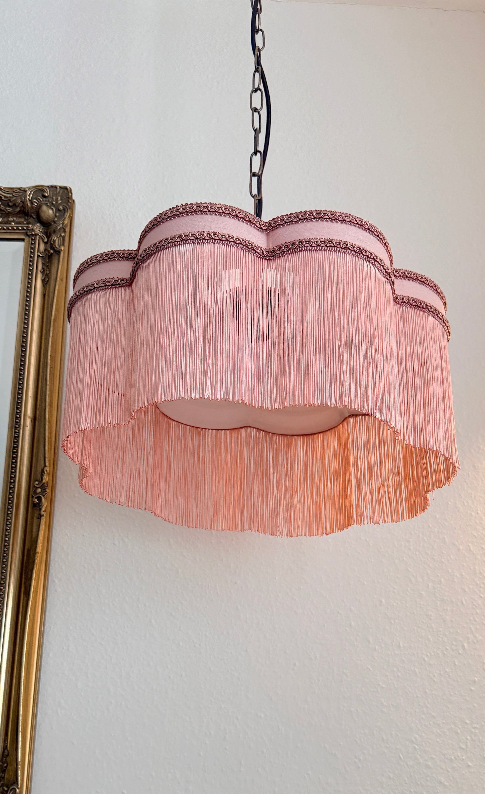 Pink fringed lampshade hanging against a white wall with a gold frame on the left.