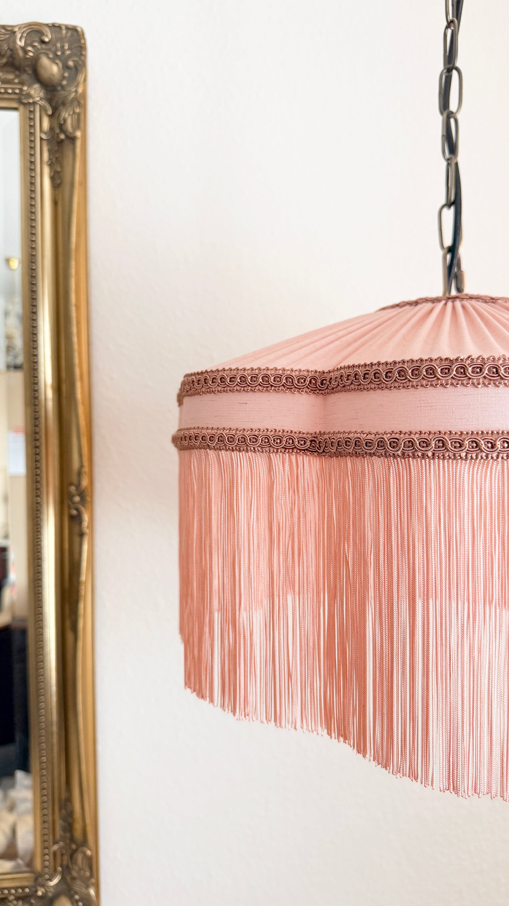 Pink lampshade with decorative trim hanging next to a gold-framed mirror on a white wall.
