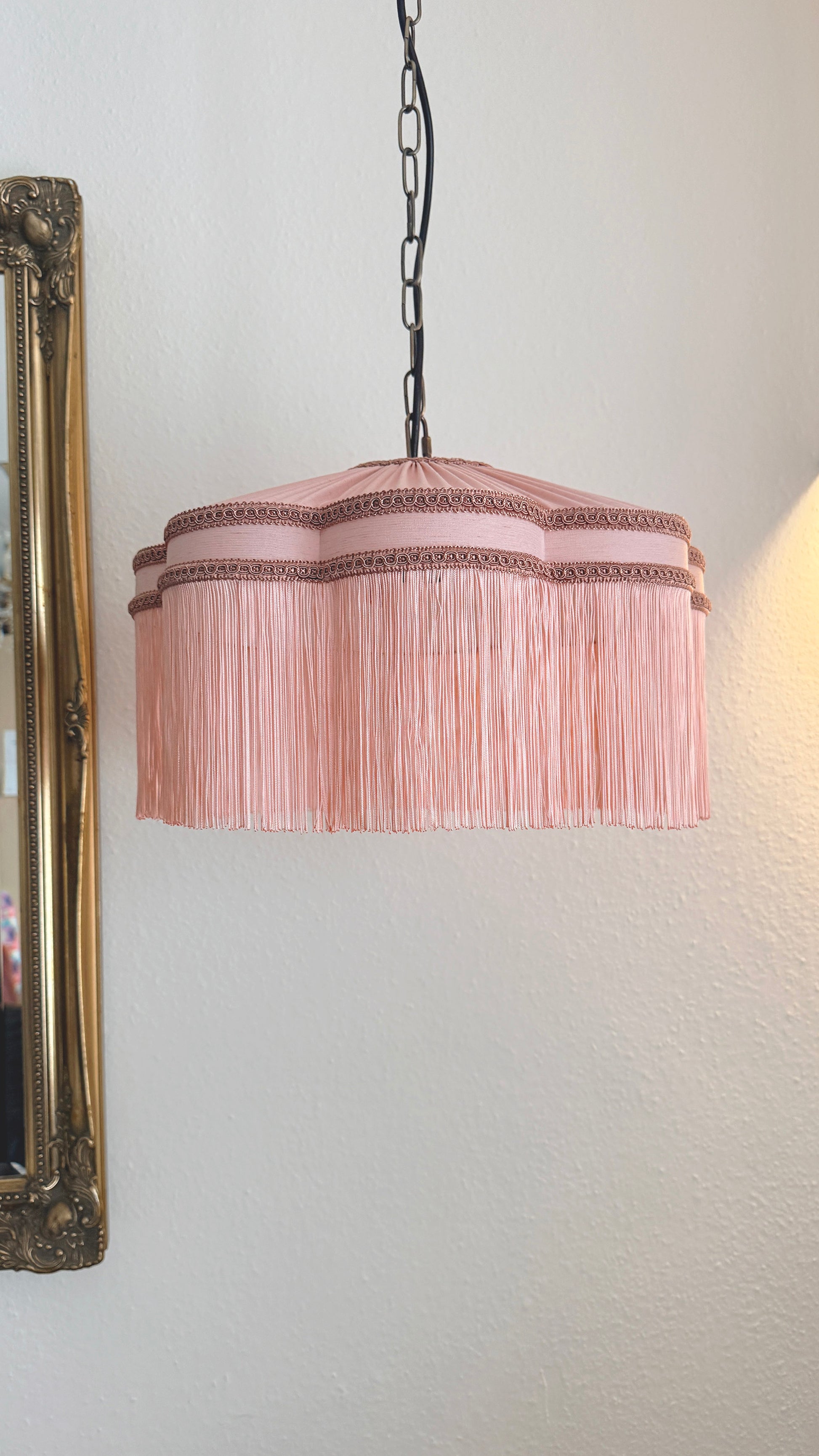 Pink pleated lampshade hanging against a white wall with a decorative mirror on the left. The House of Soft Light