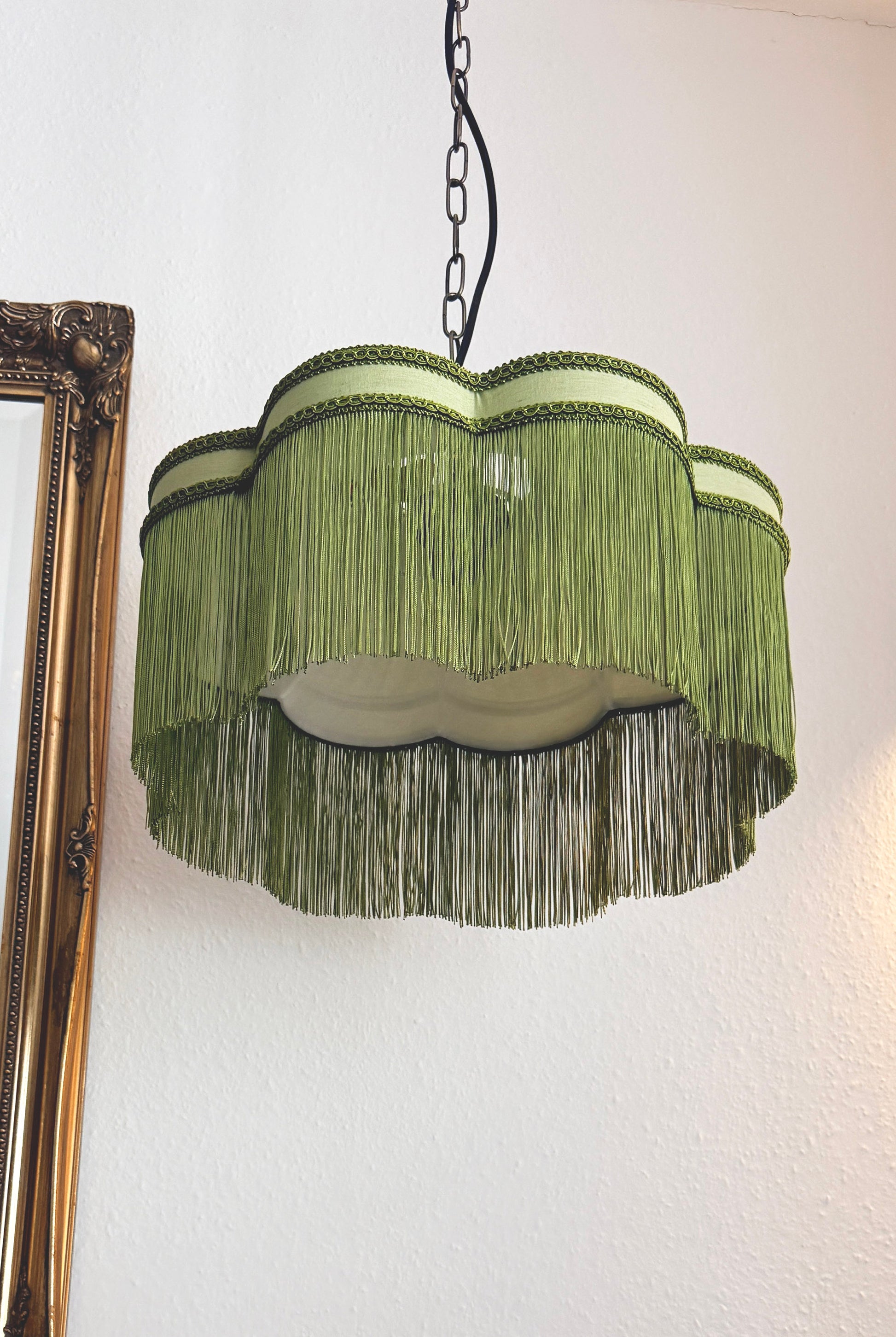 Green fringe lampshade hanging against a white wall with a gold frame on the left.