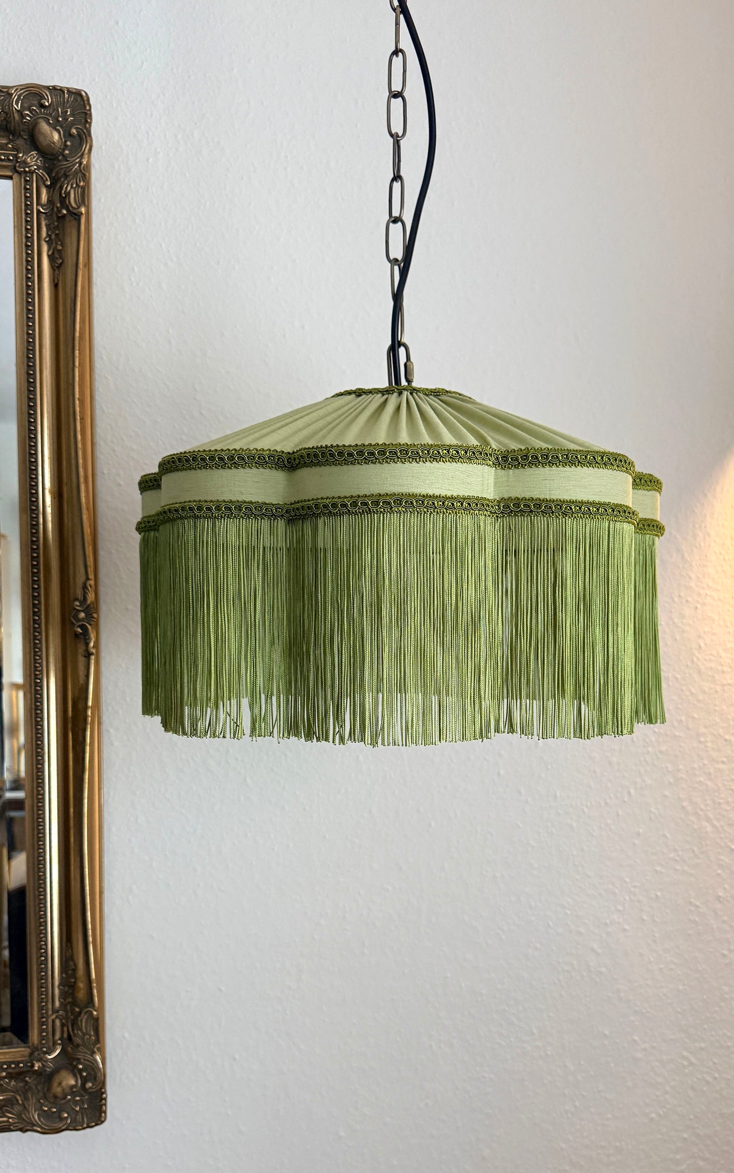 Green fringed lampshade hanging on a white wall with a decorative mirror to the left. The House of Soft Light