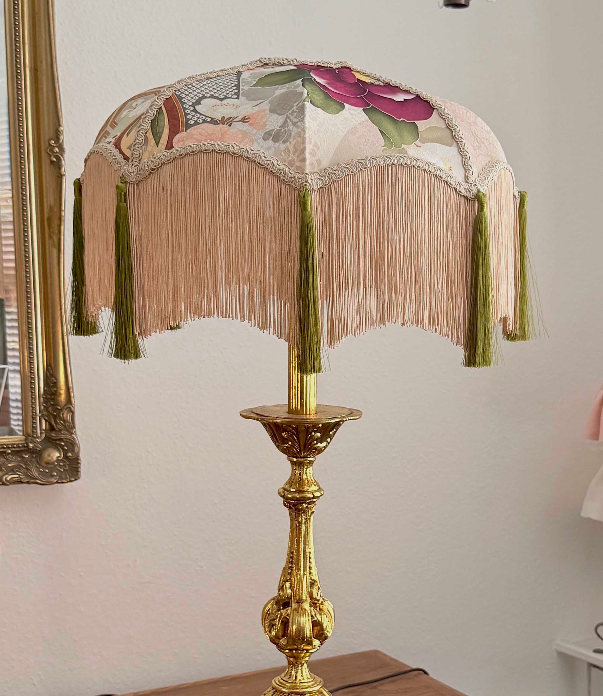 An ornate table lamp with a gold-finished pedestal base and a fringed fabric shade featuring a floral print and olive green tassels, shown against a light wall.