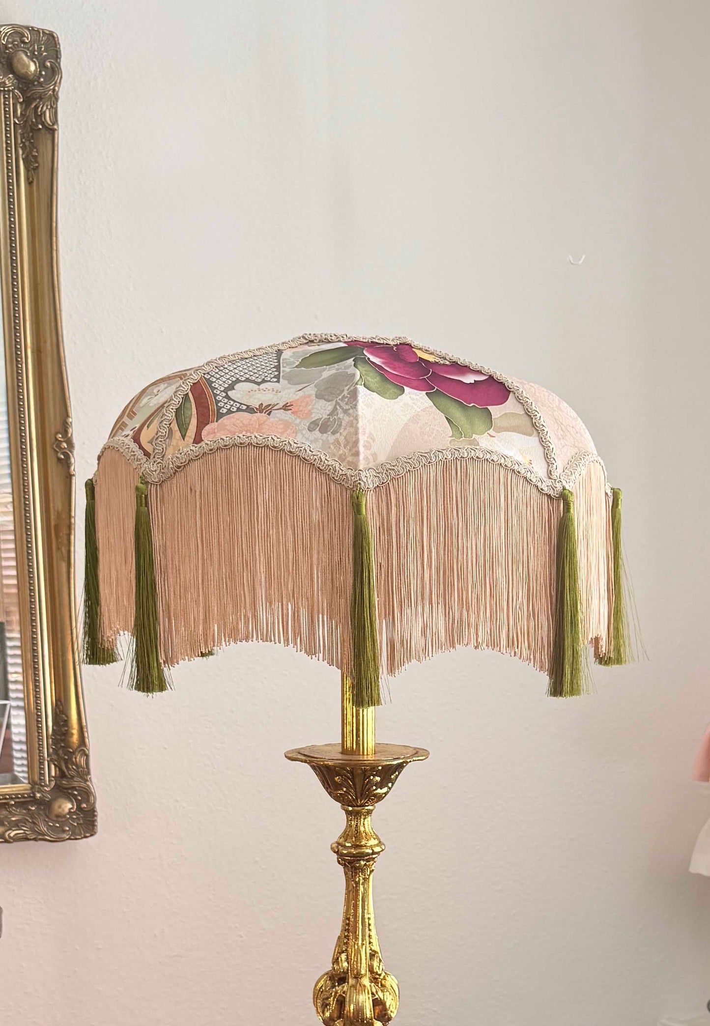 A vintage-style table lamp with a fringed fabric shade featuring floral kimono silk and a gold-toned metal base, shown against a light wall.