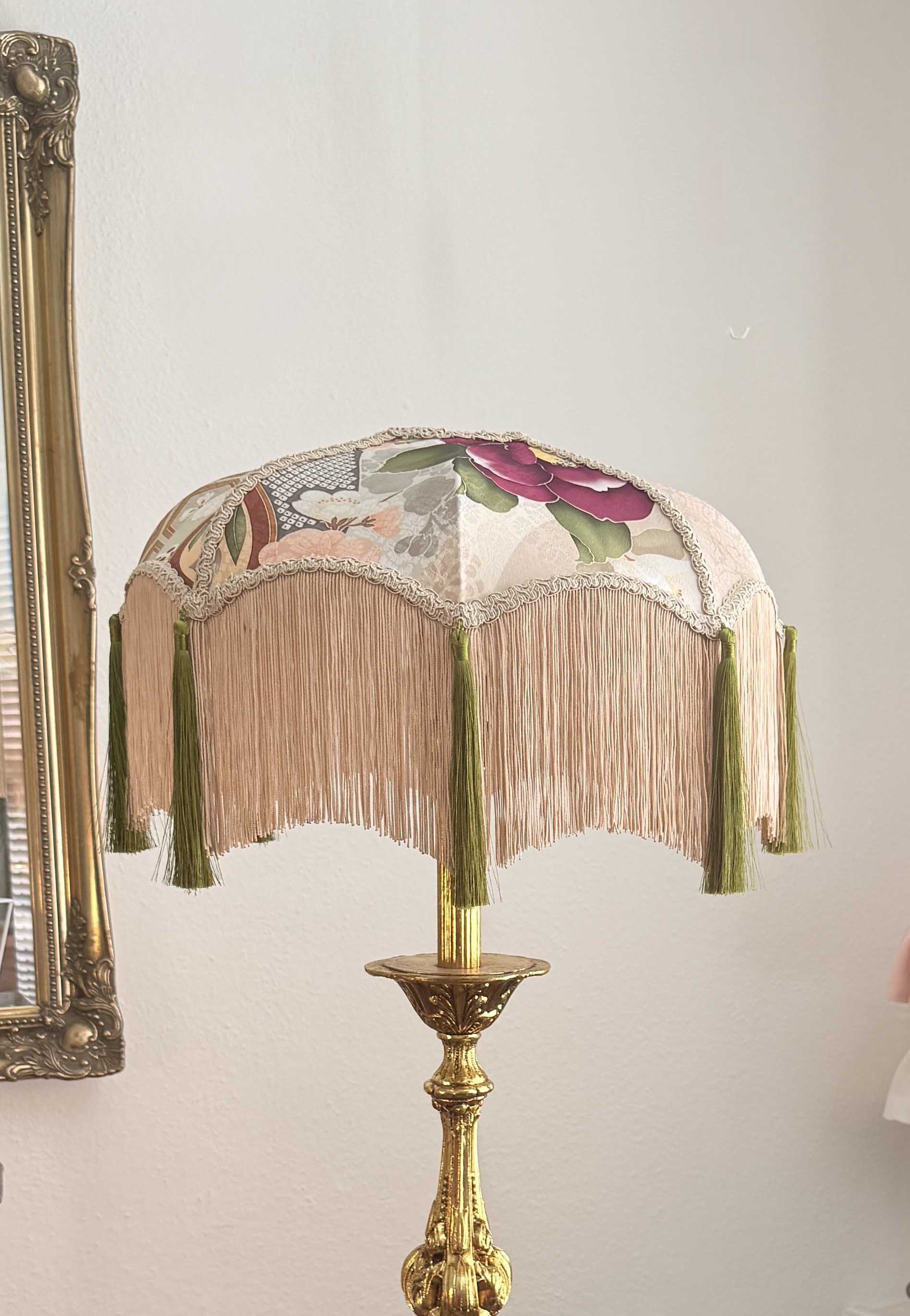 A vintage-style table lamp with a fringed fabric shade featuring floral kimono silk and a gold-toned metal base, shown against a light wall.