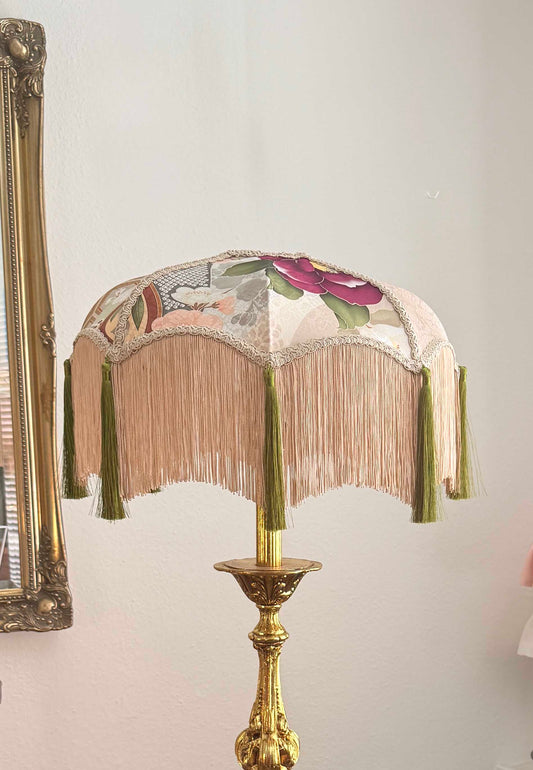 A vintage-style table lamp with a fringed fabric shade featuring floral kimono silk and a gold-toned metal base, shown against a light wall.