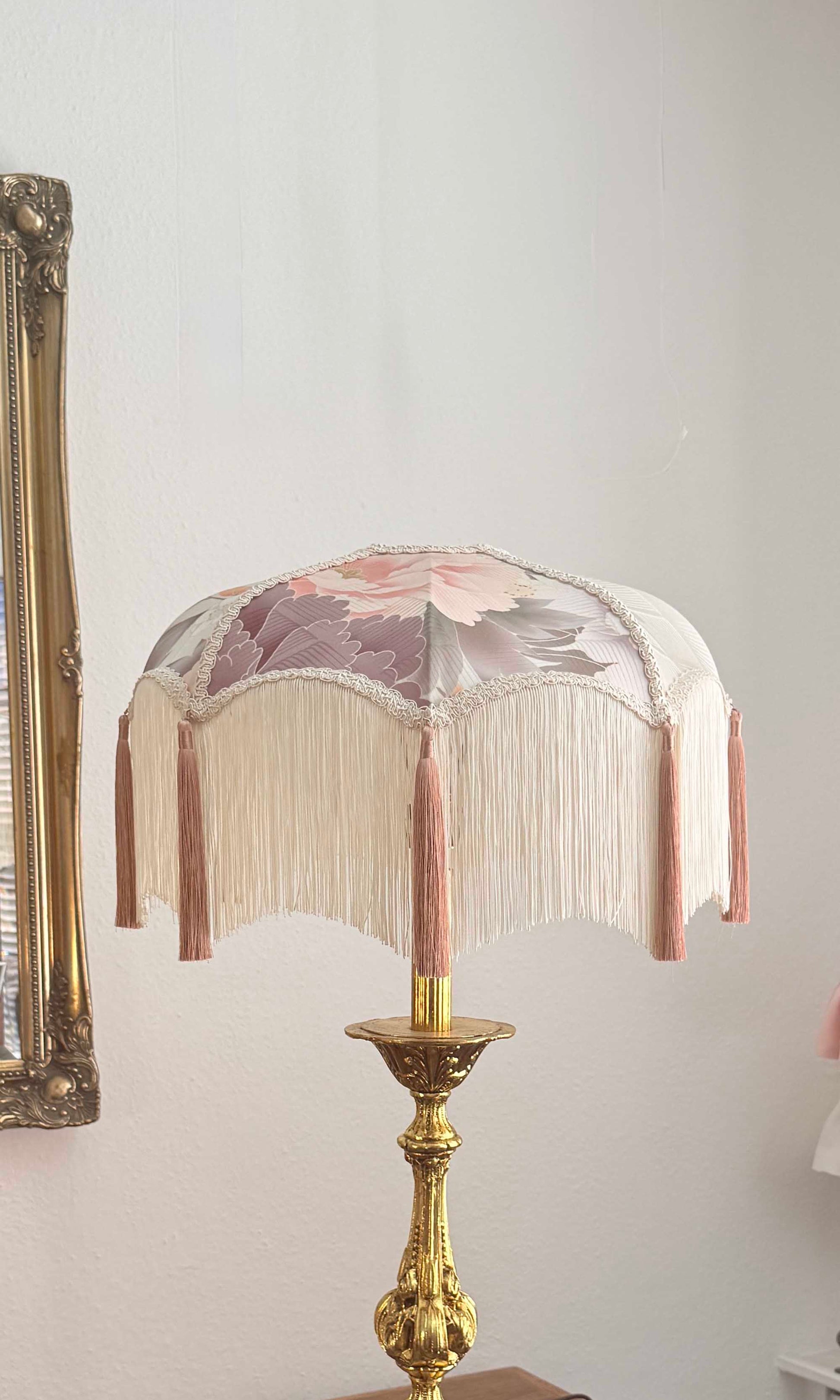 An ornate table lamp with a cream fringed shade featuring a kimono silk floral pattern in pink and mauve, mounted on a polished gold brass base.