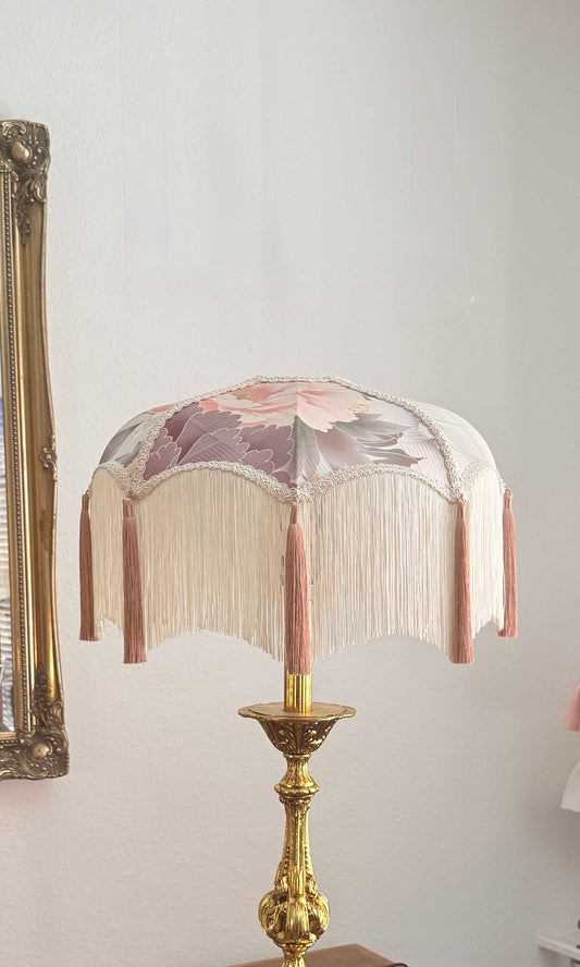 An ornate table lamp with a cream fringed shade featuring a kimono silk floral pattern in pink and mauve, mounted on a polished gold brass base.