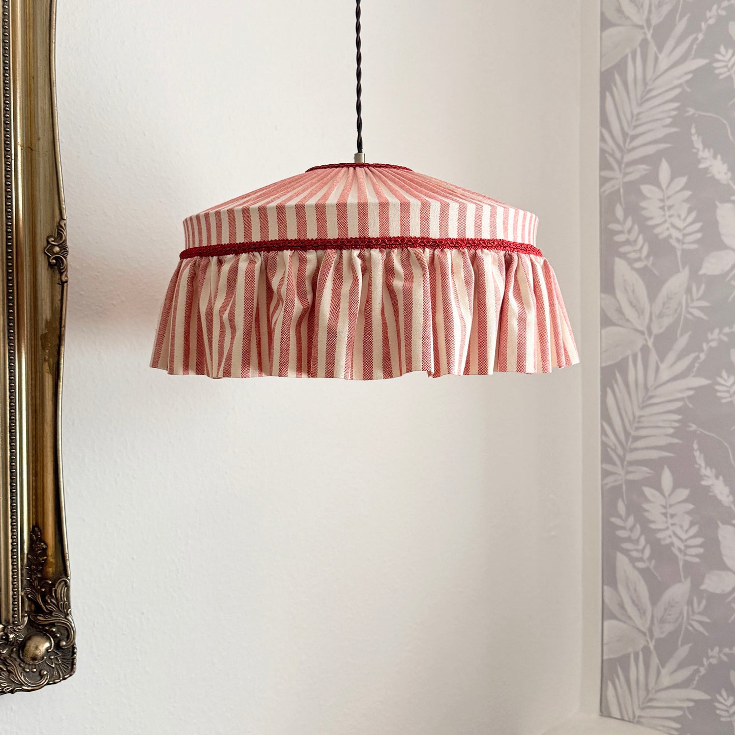 Striped lampshade hanging against a white wall with a decorative mirror and wallpaper.