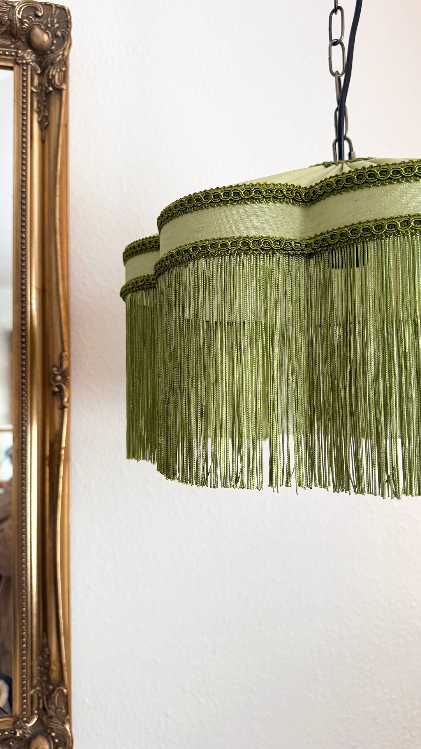 Green fringe lampshade hanging on a white wall next to an ornate mirror.