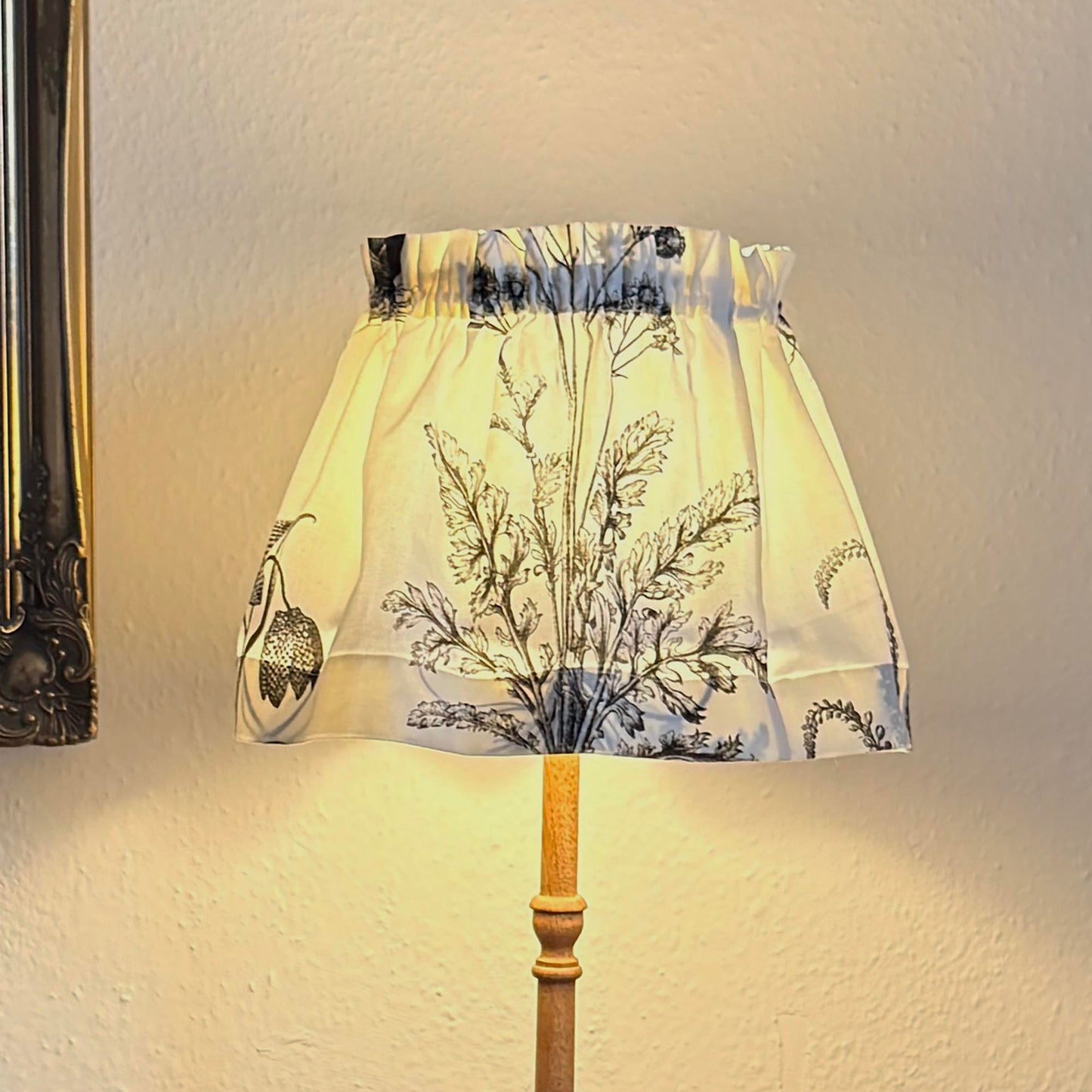 A table lamp with a cream fabric shade featuring a black botanical pattern, a light wood stem, 