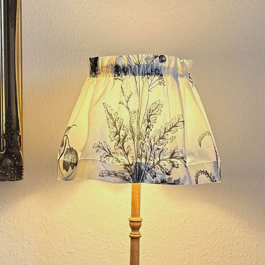 A table lamp with a cream fabric shade featuring a black botanical pattern, a light wood stem, 
