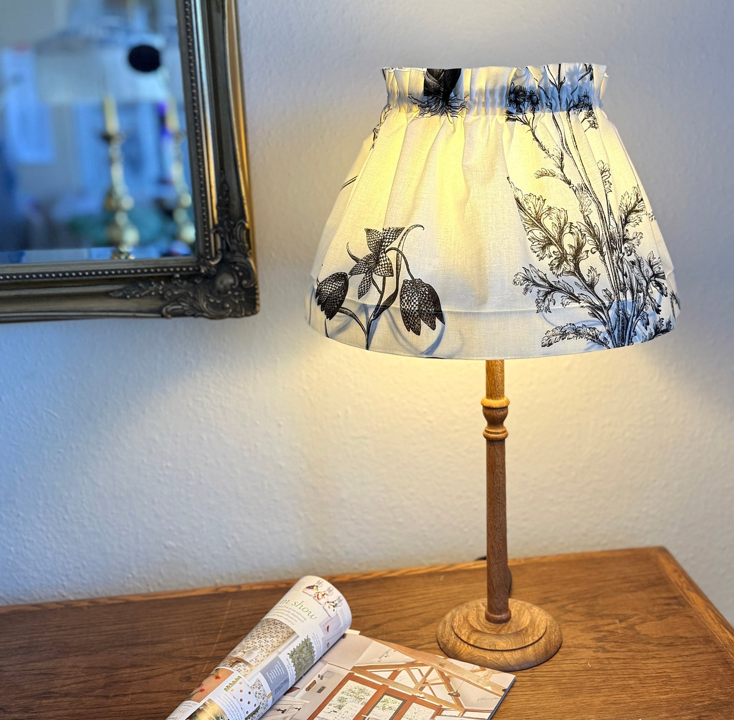 A lit table lamp with a cream fabric shade featuring a blue botanical floral pattern, set on a wooden table next to an open magazine.