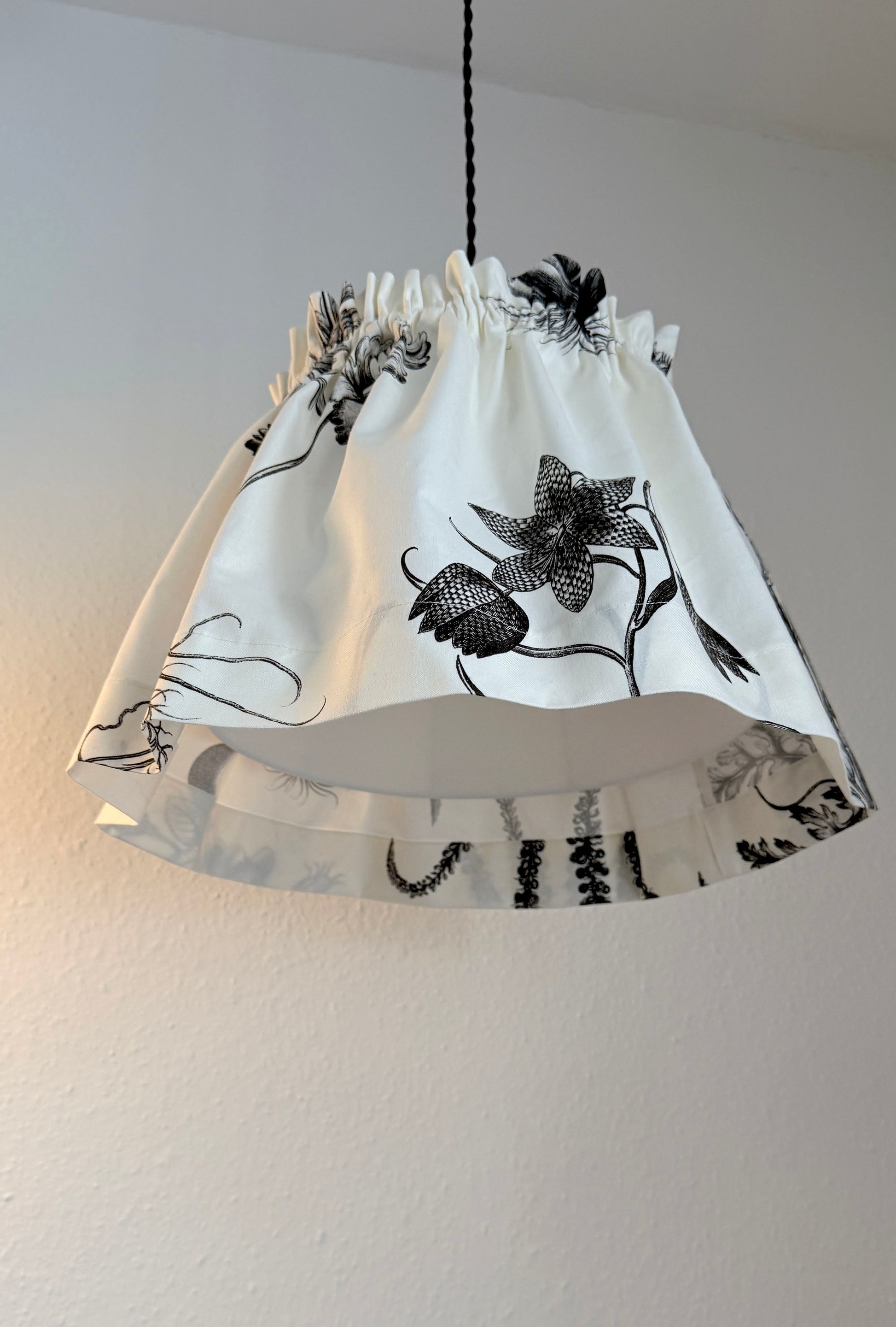 A white fabric drum shade with a black floral and botanical print, hanging from a black cord against a plain off-white wall.