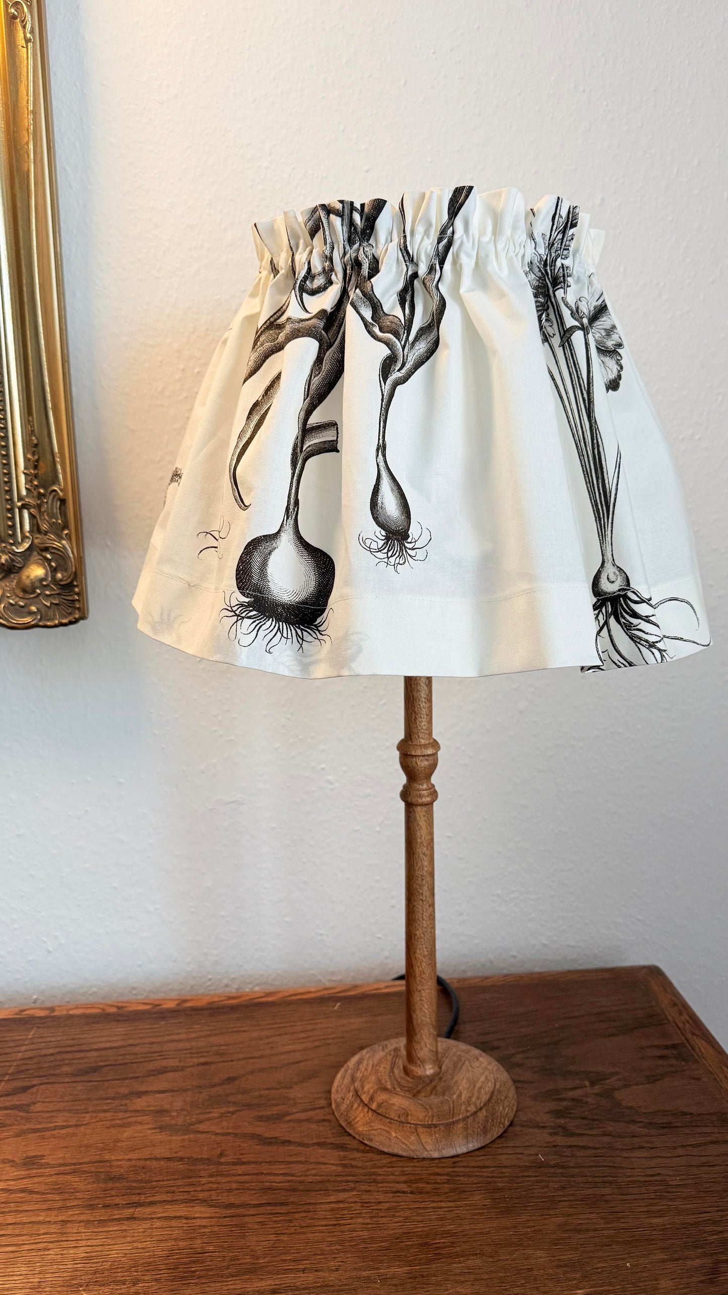 A fabric table lampshade with a black and white botanical illustration of onion bulbs, hanging from a rustic wooden lamp stand on a wooden table against a white wall.