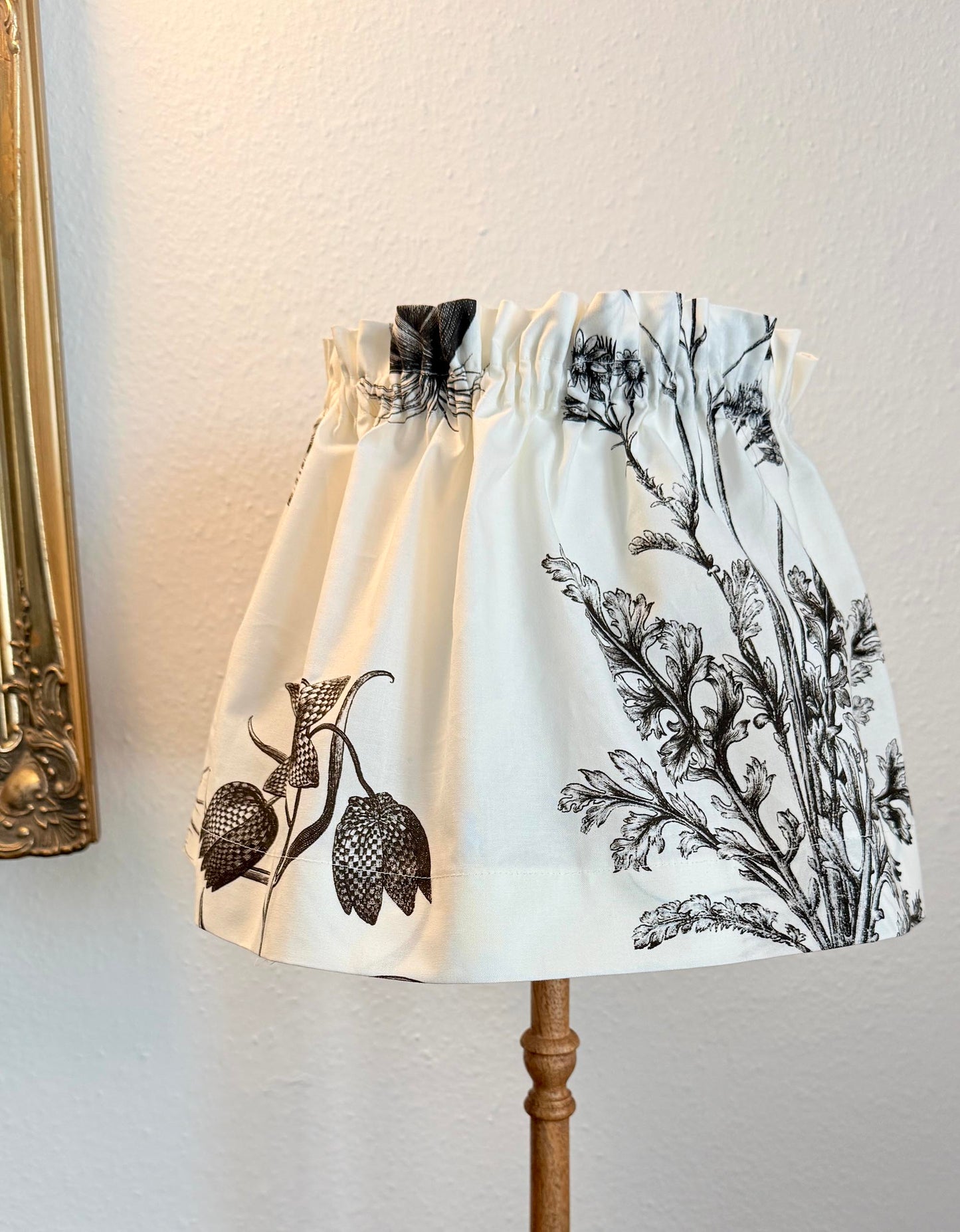 A fabric lampshade with a black botanical and floral toile-style print on an off-white background, displayed on a wooden stand against a textured cream wall.