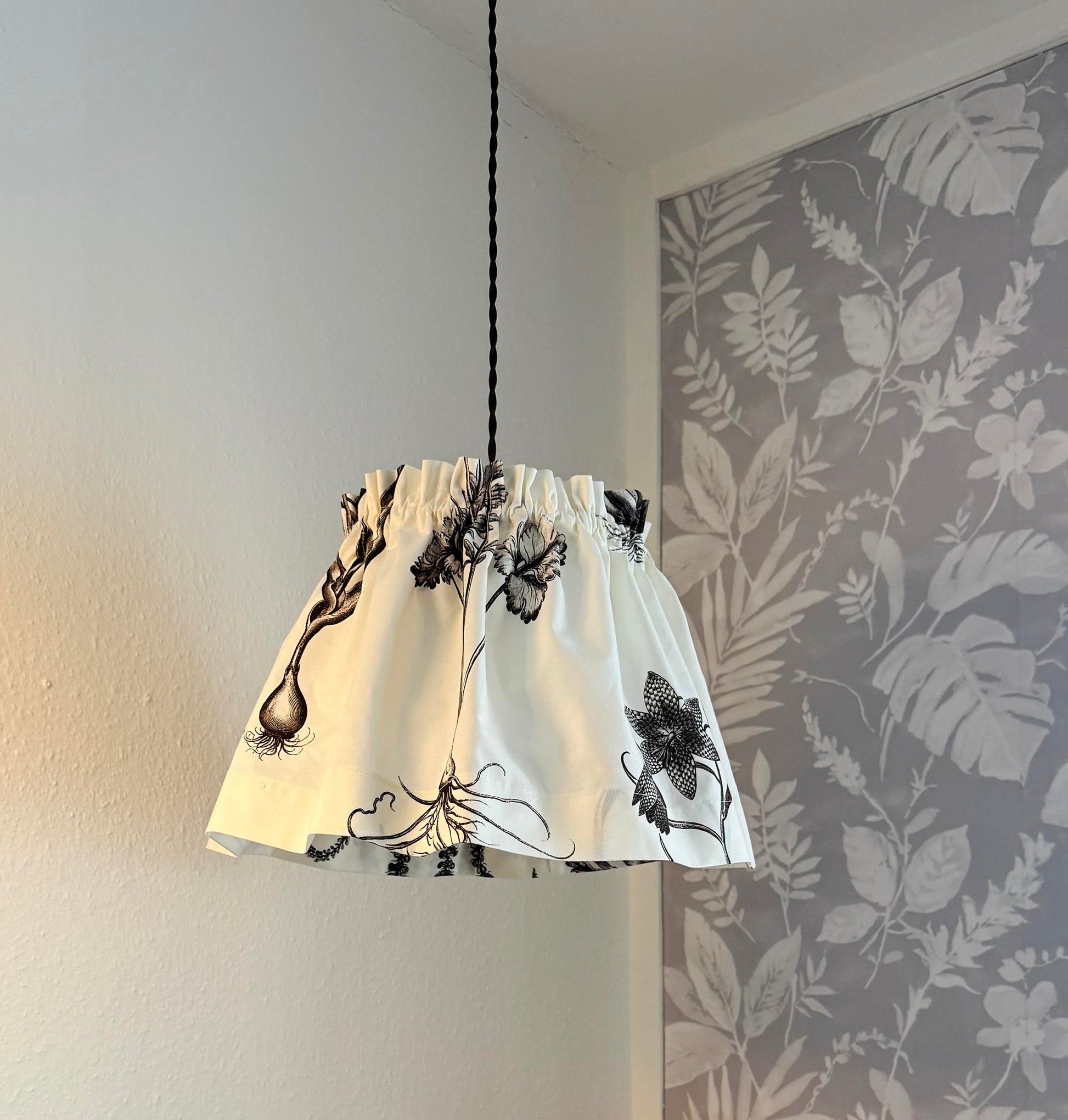 A white fabric drum pendant lampshade with a black botanical and floral print, hanging from a black cord in a room corner with a gray floral wallpapered wall.