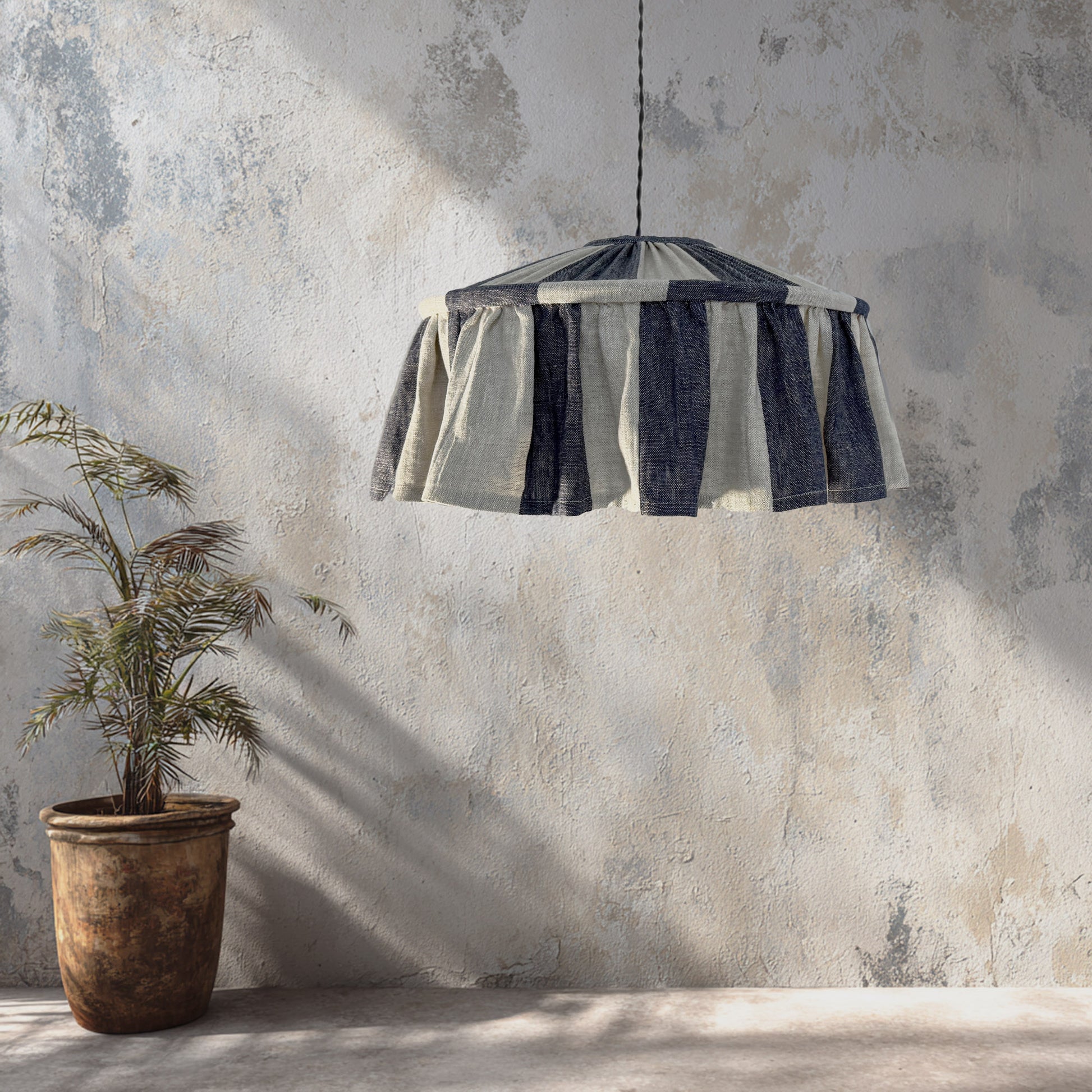 Striped lampshade hanging against a textured wall with a plant on the left.